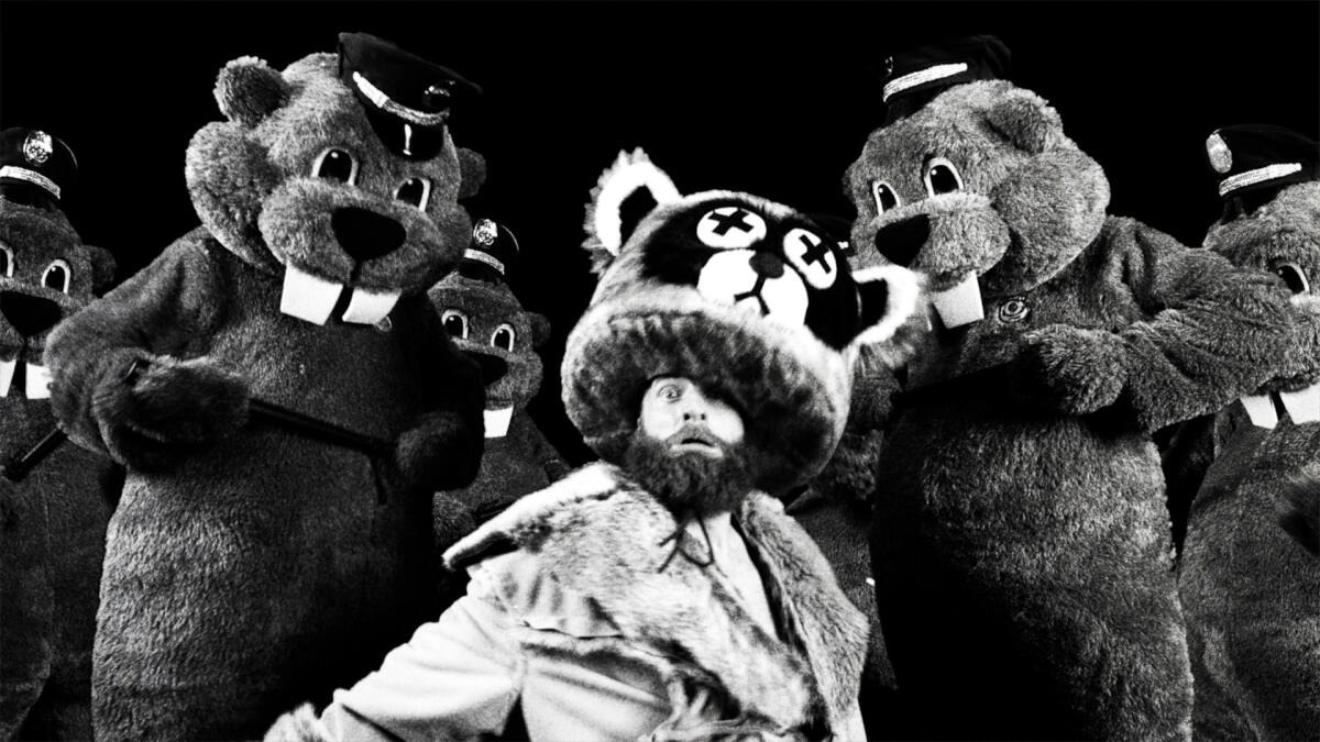 A black and white image of a bearded man wearing a raccoon animal costume. He is surrounded by two large beavers dressed as cops.