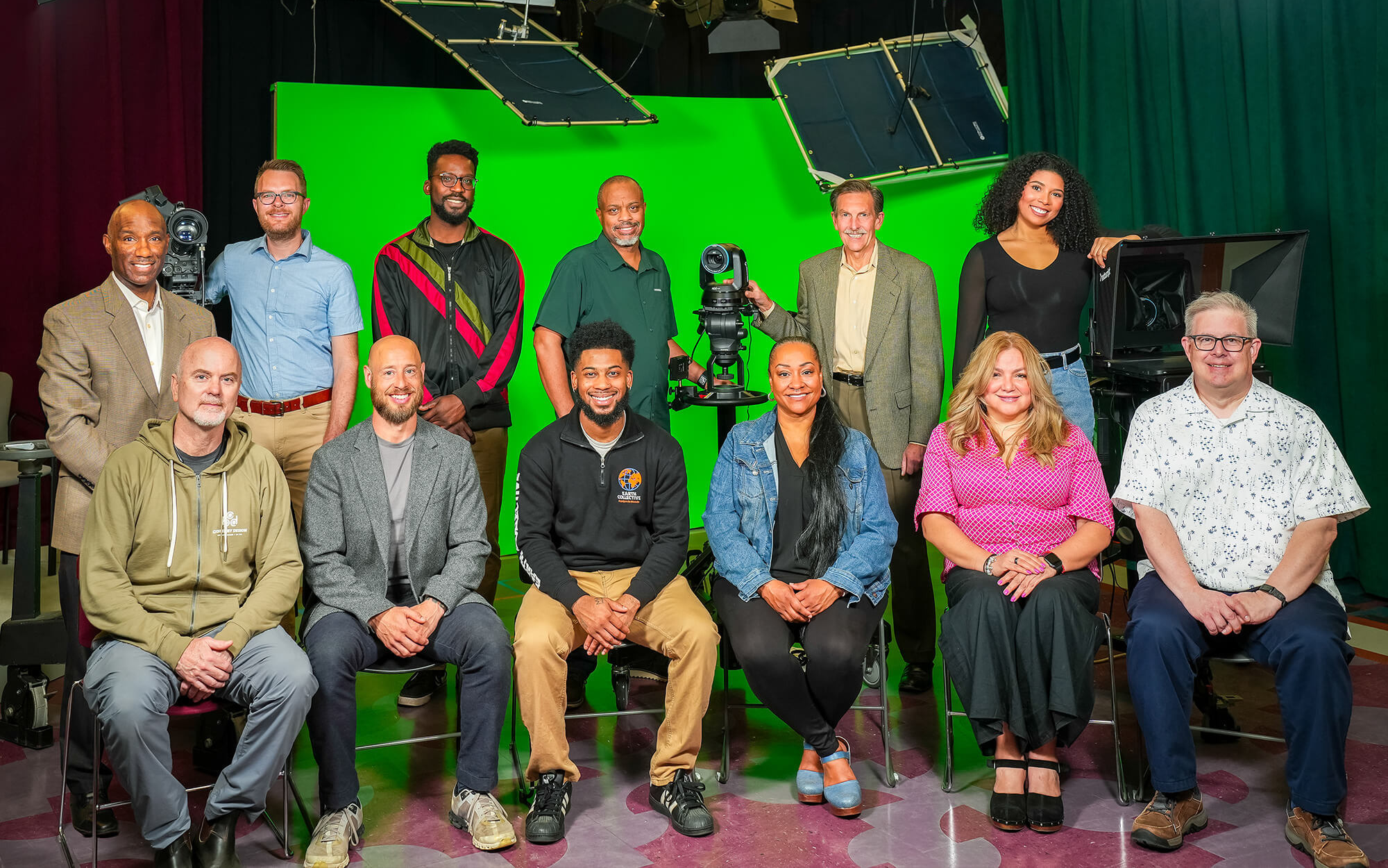The GRCMC board sits and stands in two rows of 6 people in the GRCMC studio. They are surrounded by television studio equipment, such as cameras, lights, curtains, and a green screen. The front row is sitting, the back row is standing.